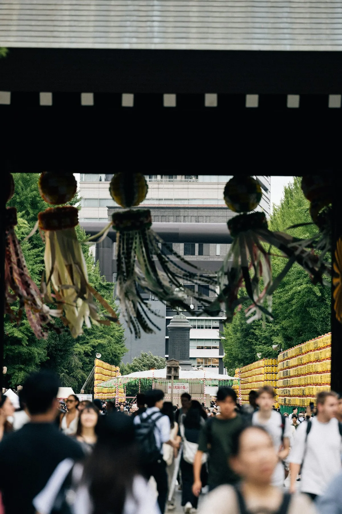 Crowds of visitors walking beneath colorful Tanabata streamer decorations hanging from a shrine gate during a Japanese summer festival, with rows of paper lanterns visible in the background