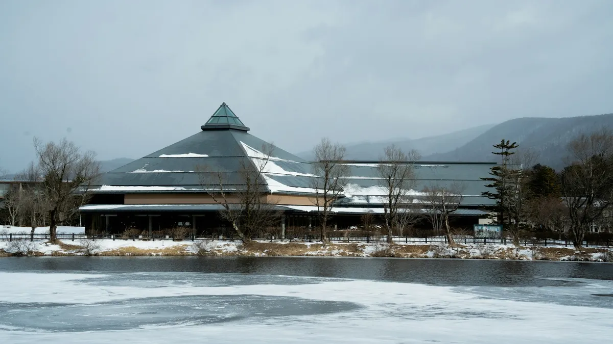 A snow-dusted building beside a partly frozen pond in Karuizawa, with bare trees and mountains behind it
