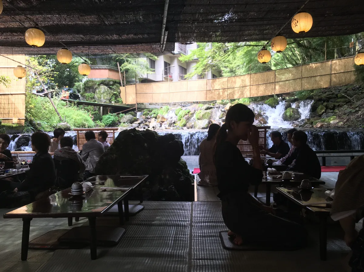 Kawadoko river dining in Kifune — guests seated on platforms over the rushing stream, with lanterns and a waterfall in the background